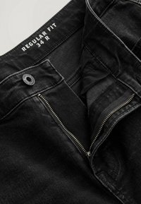Jean en denim noir avec braguette non fermée, bouton en métal et étiquette "Regular Fit 34 R" sur la ceinture intérieure.