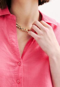 Gouden ketting met een gladde schakels, gedragen over een roze linnen shirt met een button-down kraag en zichtbare knoopdetails.