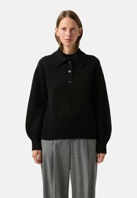 Black knitted pullover with a large collar and three-button placket; features wide sleeves and a ribbed hem, styled with gray pleated trousers.