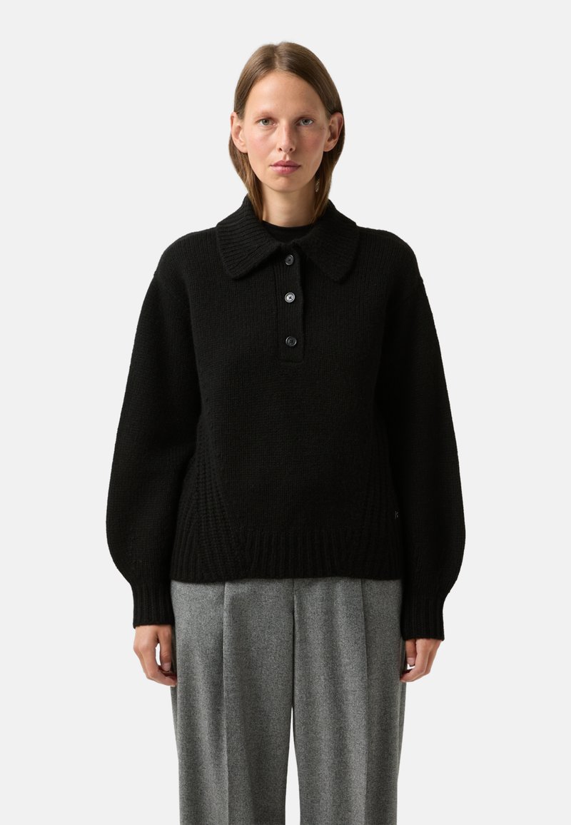 Black knitted pullover with a large collar and three-button placket; features wide sleeves and a ribbed hem, styled with gray pleated trousers.