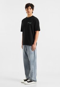 Svart bomulls T-skjorte med korte ermer som har en subtil blå grafikk. Brukt med lyseblå baggy jeans og svart-hvite sneakers.