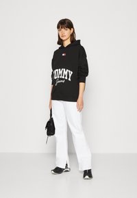 Felpa nera con la scritta "TOMMY Jeans" in bianco, caratterizzata da un piccolo logo applicato. Abbinata a pantaloni bianchi e sneaker nere. Design semplice.