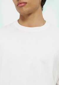 Νεαρό άτομο που φοράει ένα απλό λευκό T-shirt με στρογγυλή λαιμόκοψη και λεπτή ασημένια αλυσίδα στον λαιμό, εμφανιζόμενο από το λαιμό έως το πάνω μέρος του στήθους.