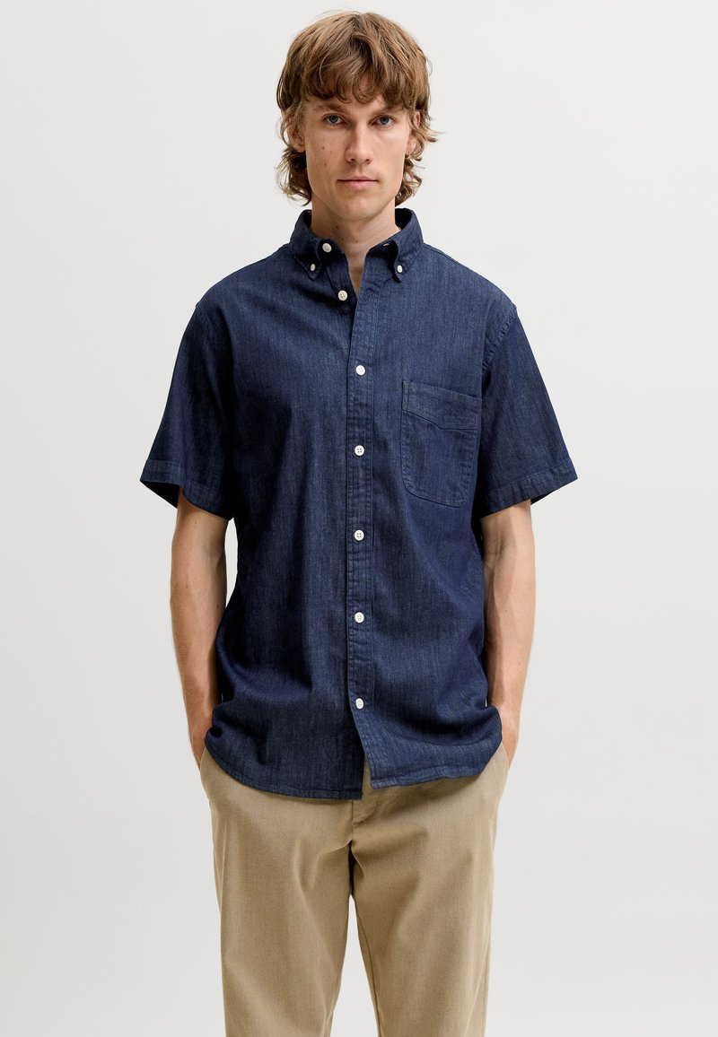 Jeune homme aux cheveux châtain clair portant une chemise bleu foncé à manches courtes boutonnée et un pantalon beige, debout les mains dans les poches.