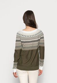 Pull en tricot avec un large col blanc, orné de motifs géométriques verts, beiges et noirs. Manches longues avec poignets côtelés.