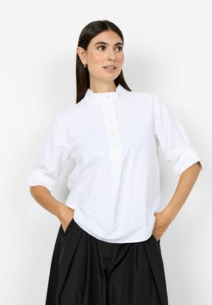 Frau mit langen dunklen Haaren, die ein weißes, geknöpftes Hemd mit hochgekrempelten Ärmeln und eine lockere schwarze Hose trägt, Hände in den Taschen, schaut seitlich.