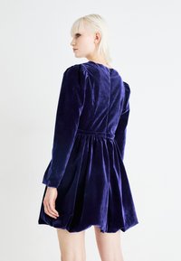 Purppuran samettimekko, jossa on pitkät hihat, puffattuja olkapäitä, kapea vyötärö ja leveä helma; takana avaus, jossa on avainaukko.