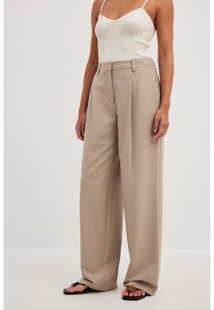 Frau in beigeweiten Hosen, einem weißen, gerippten Spaghettiträgertop und schwarzen Sandalen mit dekorativen Details.