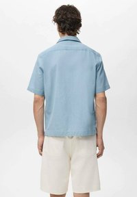 Camisa de manga corta en algodón azul claro, con un corte holgado y cuello, combinada con pantalones cortos de color crema y costuras traseras visibles.