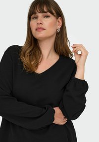 Crna dugorukava majica s V-izrezom, teksturiranim materijalom i naboranim manžetama. Model pozira s podignutom rukom i mekim valovima u kosi.