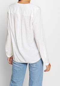 Weiße Bluse mit langen Ärmeln und Knopfmanschetten, mit kleinen schwarzen Punkten und einem Rückeneinsatz. Glattes Material, lockere Passform.