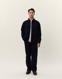 Completo in velluto a coste navy con motivo floreale, dotato di design con bottoni e una t-shirt beige sotto. Scarpe marroni.