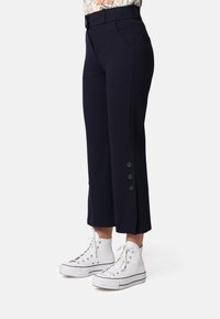 Pantalones de talle alto en color marino con perneras recortadas, que presentan un cierre lateral, presillas para cinturón y tres botones negros en el dobladillo del tobillo. Combinados con zapatillas blancas.