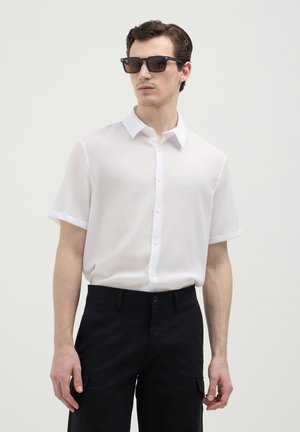 Camicia bianca a maniche corte con tessuto testurizzato. Abbinata a pantaloni cargo neri e occhiali da sole rettangolari.