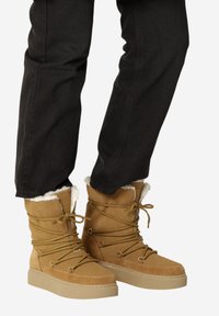 Botas de tobillo de ante color topo con un forro blanco suave, suelas planas beige y cordones decorativos. Textura suave y diseño elegante con toques metálicos.