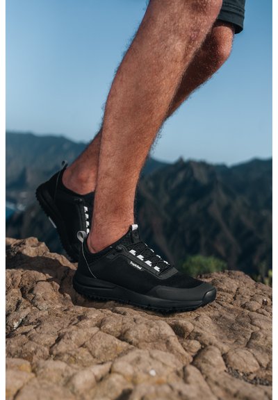 Black athletic shoes with a mesh upper, white laces, and textured soles, set against a rocky landscape.