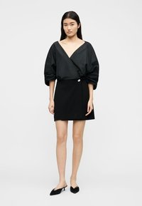 Jeune femme aux longs cheveux noirs portant un chemisier noir à col en V profond avec des manches bouffantes, une jupe portefeuille noire et des mules noires pointues.