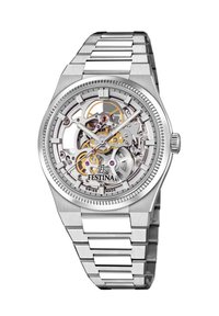 Reloj de acero inoxidable plateado con una esfera esqueletizada transparente que muestra engranajes dorados y joyas moradas, con marcadores horarias luminosos.