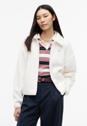 Donna con lunghi capelli scuri indossa una giacca in pile bianca sopra una polo a righe rosse, bianche e blu e pantaloni blu navy a righe sottili con cintura marrone.
