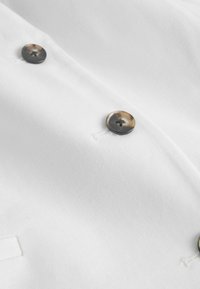 Tissu blanc avec trois boutons en écaille cousus en ligne verticale, deux boutonnières visibles sur le côté gauche.