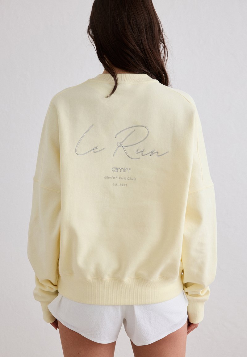 Frau mit langen dunklen Haaren trägt ein blassgelbes Sweatshirt mit dem Schriftzug "Le Run" auf dem Rücken und weiße Shorts, steht vor einem hellen Hintergrund.
