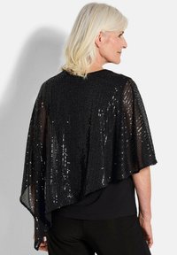 Schwarze Pailletten-Cape mit lockerer Passform, abgerundetem Rücken und strukturiertem Oberflächen, über einem schwarzen Top geschichtet.