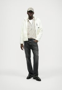 Veste zippée en polaire blanche sur un haut beige, jeans gris et chaussures noires, complétée par une casquette gris clair. Design simple et décontracté.