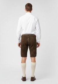 Pantaloni corti in pelle marrone con finitura testurizzata, abbinati a una camicia bianca a maniche lunghe, calze color crema al ginocchio e scarpe marroni.