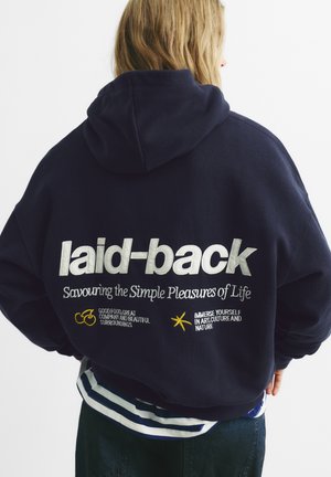 Marineblå hættetrøje med stort broderet tekst på ryggen: "laid-back" og "Savouring the Simple Pleasures of Life" med farverige ikoner.