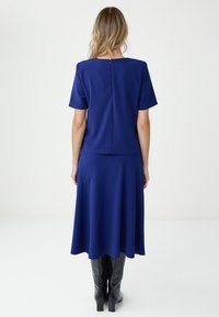 Kobieta o blond włosach ubrana w niebieski top z krótkim rękawem, pasującą midi spódnicę i czarne kozaki do kolan, stoi tyłem na białym tle.