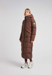 Abrigo acolchado marrón con capucha, con un diseño largo y acolchado, cremallera frontal y bolsillos laterales. Llevado con zapatillas blancas y plateadas.