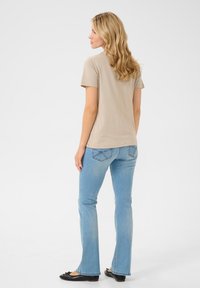 Beige kortærmet T-shirt kombineret med lyseblå klokkeformede jeans med baglommer. Modellen står med hår, der falder ned over skuldrene.