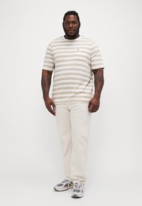Beige és fehér csíkos, rövid ujjú póló zsebbel, amely off-white nadrággal és szürke sportcipővel van párosítva. Lapos anyag, laza szabású.