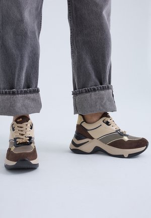 Persona con vaqueros grises remangados y zapatillas gruesas beige y marrón con detalle de cordones, de pie sobre una superficie gris clara.