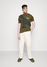 Olivegrünes T-Shirt mit kurzen Ärmeln und Logo, crèmefarbene weit geschnittene Hose und grün-schwarze Sneakers. Grauer Kapuzenpullover über den Schultern drapiert.