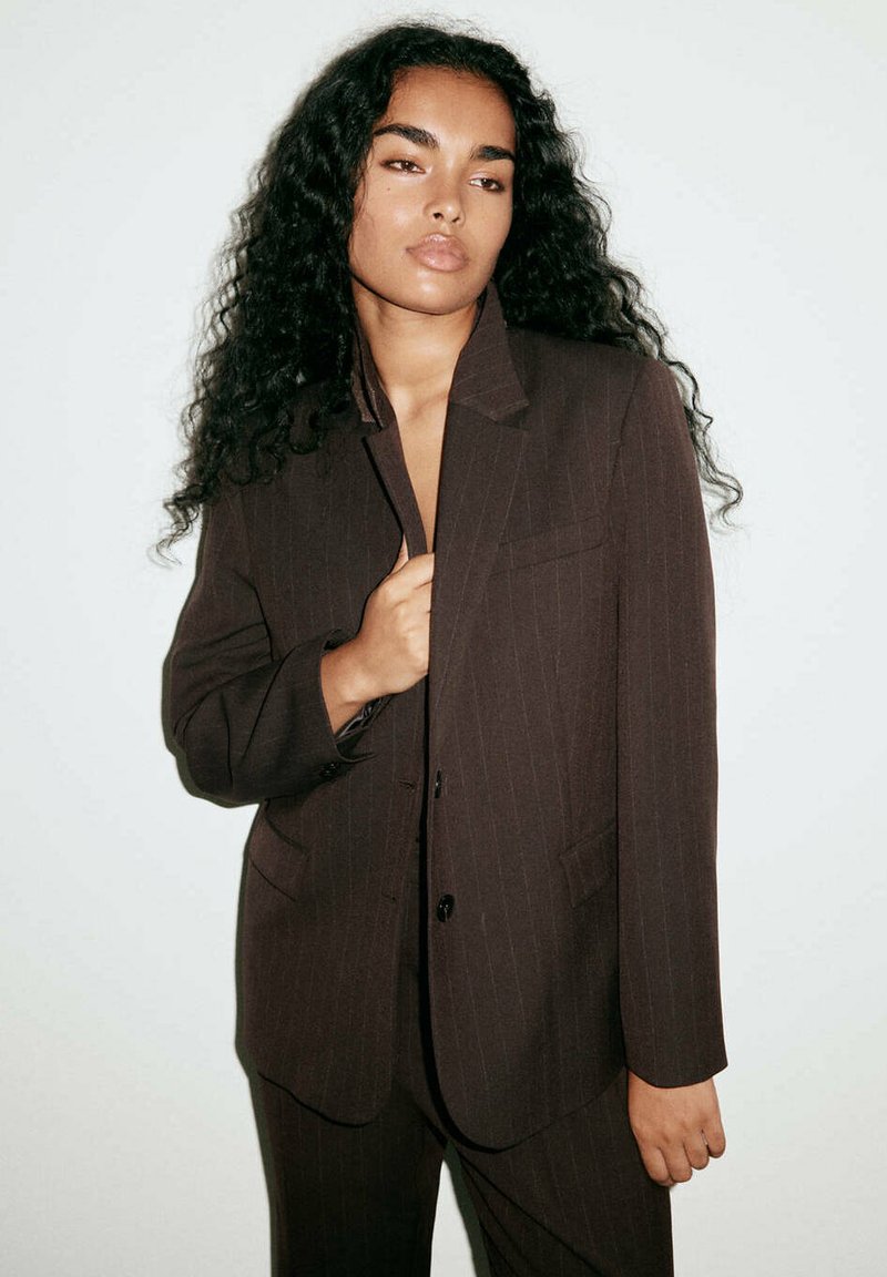 Femme aux longs cheveux bouclés portant une veste de costume marron foncé à fines rayures, tenant le revers d'une main sur un fond clair uni.