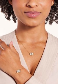 Gouden ketting en armband met een centraal verweven knoopontwerp, voorzien van een heldere edelsteenaccent op elk stuk.