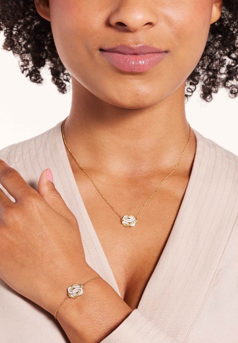 Gouden ketting en armband met een centraal verweven knoopontwerp, voorzien van een heldere edelsteenaccent op elk stuk.