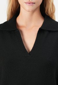 Close-up de uma mulher a usar um suéter de malha preto com um decote em V profundo e gola larga, a mostrar o seu pescoço e lábios.