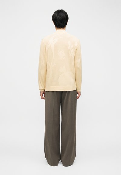 Άτομο που φοράει μπεζ πουκάμισο με μακριά μανίκια και διακριτικά λουλουδενια σχέδια, καθώς και χαλαρά γκρι παντελόνια, στέκεται γυρισμένο με την πλάτη σε ένα απλό φόντο.