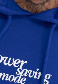 Felpa con cappuccio blu realizzata in morbido tessuto, con testo bianco che recita "modalità risparmio energetico". Sono visibili i lacci e un primo piano del collo.