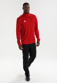 Rød sportslig zip-up topp med hvite detaljer, kombinert med svarte bukser med hvite striper. Materialet ser lett og pustende ut.