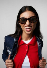 Occhiali da sole tartarugati con montatura ovale oversize, indossati con una camicia rossa con bottoni sotto un giubbotto di denim. Texture liscia, design casual.