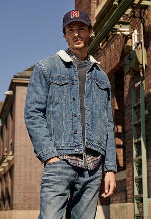 Veste en denim avec un col en shearling, de couleur bleue, deux poches poitrine, portée sur un pull gris et une chemise à carreaux, associée à un jean bleu et une casquette.