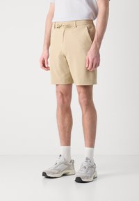 Pantalones cortos beiges con textura, cintura con cordón, bolsillos laterales y un corte relajado, combinados con zapatillas gris claro y calcetines blancos.
