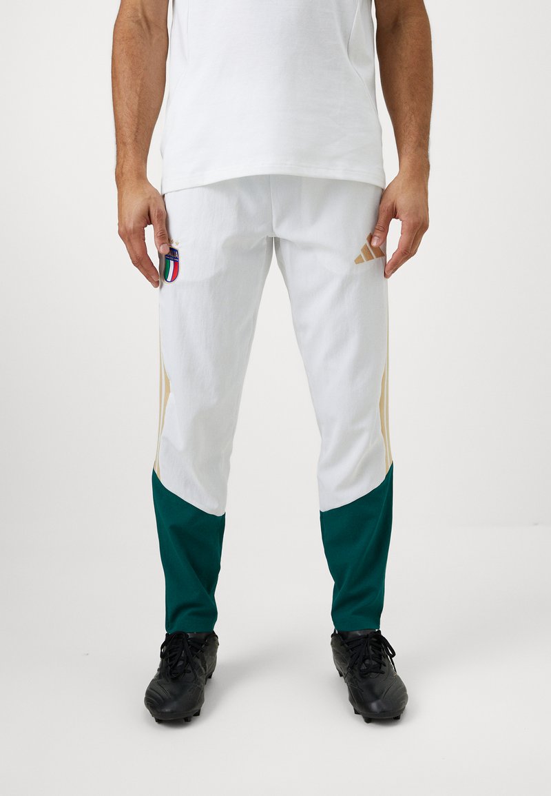 Άνδρας φορώντας άσπρο και πράσινο αθλητικό παντελόνι με το έμβλημα της Ιταλίας και το λογότυπο της Adidas, σε συνδυασμό με μαύρες παπούτσια, στέκεται μπροστά σε απλό φόντο.