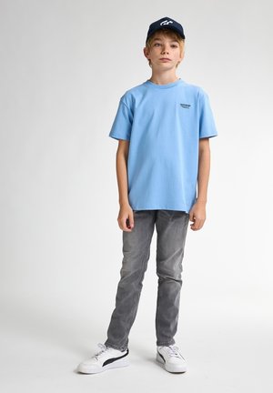 Un tricou din bumbac de culoare albastru deschis, cu mânecă scurtă și un mic logo negru, completat cu blugi gri și adidași albi cu accente negre.