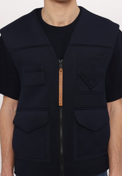 Gilet zippé bleu marine avec une texture lisse, doté de deux poches avant, d'une tirette en cuir et d'un logo sur la poche supérieure.