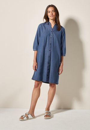 Robe chemise en denim avec un devant boutonné, des manches trois-quarts et une coupe décontractée. Présente une texture subtile et une longueur mi-mollet. Associée à des sandales en argent.