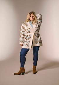 Ženska v vzorčastem kardiganu s pasom, svetlo modrih majici, temno modrih kavbojkah in rjavih gležnjarjih, pozira z roko na glavi.